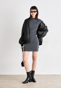 Γκρι εφαρμοστό φόρεμα συνδυασμένο με μια μαύρη δερμάτινη τζάκετ bomber, μαύρα μυτερά μποτάκια και πράσινα γυαλιά ηλίου, κρατώντας μια μικρή τσάντα.