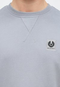 Lys grå sweatshirt med rund hals, korsstingsdetaljer på kragen og en firkantet logopatch med et stilisert emblem.