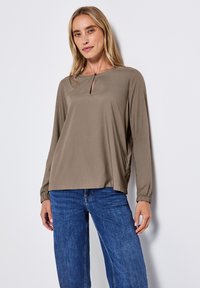Blusa a maniche lunghe in taupe con scollatura rotonda e dettaglio a chiave sul davanti. Presenta polsini elastici e una vestibilità rilassata. Abbinata a jeans blu.