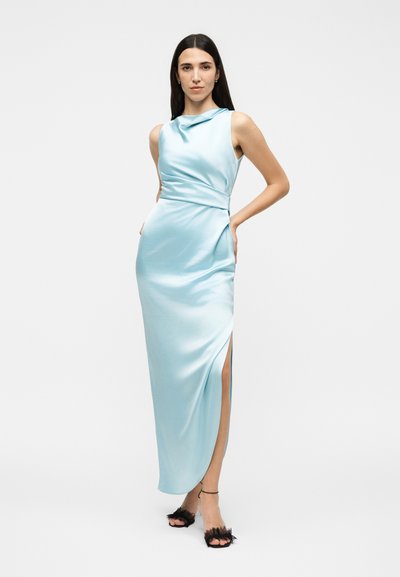 Γυναίκα με αμάνικο ανοιχτό γαλάζιο σατέν φόρεμα με σκίσιμο στο πλάι, στέκεται με το ένα χέρι στη μέση, φοράει μαύρα σανδάλια με ψηλό τακούνι και λουράκια στον αστράγαλο.