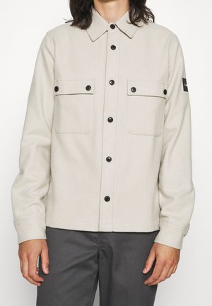 Beige skjortejakke med to brystlommer og sorte knapper, båret af en person med mørkegrå bukser, stående mod en hvid baggrund.