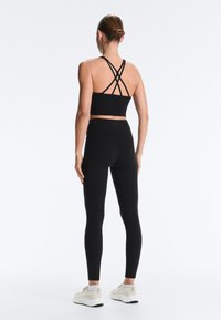 Žena v černém krátkém topu s křížovými ramínky a černých legínách s vysokým pasem, stojící zády, obutá v bílých sportovních botách na bílém pozadí.