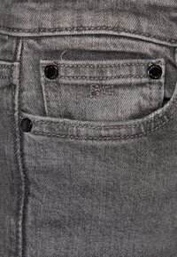 Harmaa farkkukangas, jossa on hieman tekstuuria. Kankaassa on pieni etutasku, jossa on mustat metalliset nupit ja sävy-sävyyn ompelu.