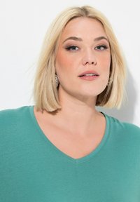 Teal V-Ausschnitt-Shirt aus weichem Stoff mit kurzen Ärmeln. Das Model hat blonde Haare und trägt runde silberne Ohrringe.