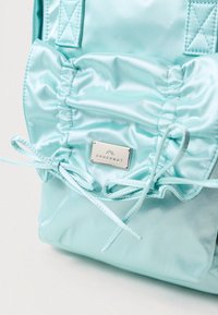 Svijetloplava ruksak izrađen od satena ima nabranu teksturu, zatvaranje na vezicu i srebrnu metalnu oznaku s dizajnom planine.