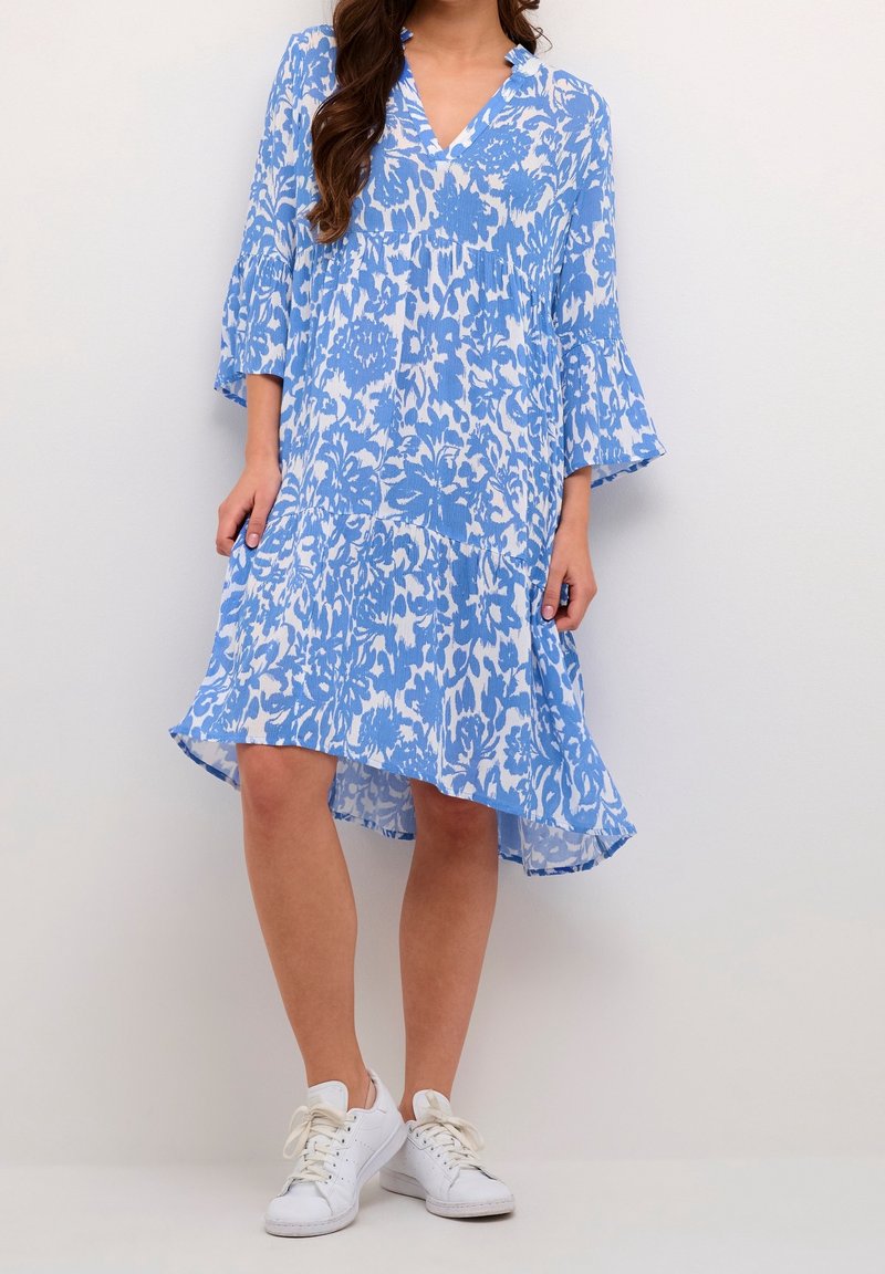 Femme portant une robe bleu et blanc à fleurs, longueur genou, avec des manches cloches et des baskets blanches, debout devant un fond uni.