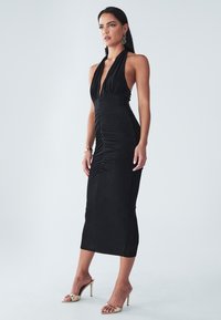 Crna halter haljina s prilagođenim dizajnom, nabrano pojačanje na prednjem delu i elegantna silueta, uparena sa zlatnim sandalama na otvorene nožice.