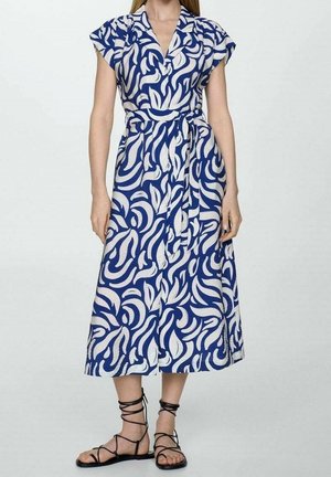 Femme portant une robe portefeuille à imprimé abstrait bleu et blanc avec des manches courtes et des sandales plates noires à lacets, debout devant un fond clair uni.