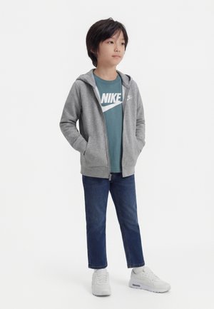 Niño con sudadera gris con cremallera, camiseta Nike turquesa, vaqueros azul oscuro y zapatillas blancas, de pie con las manos en los bolsillos.