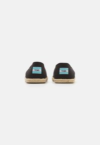 Des chaussures slip-on en toile noire avec une semelle en jute, arborant des étiquettes TOMS turquoise sur le talon et un design minimaliste.
