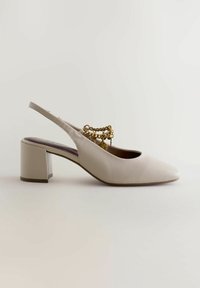 Chaussure à talon bloc beige à bride arrière, avec bout pointu et chaîne décorative dorée sur le dessus, présentée de profil sur un fond uni.