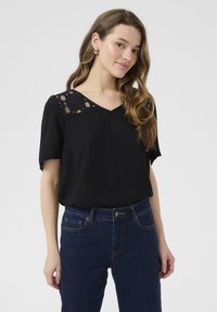 Blouse noire à manches courtes, décolleté en V et détails en dentelle aux épaules. Portée avec un jean en denim bleu foncé, texture lisse et coupe décontractée.
