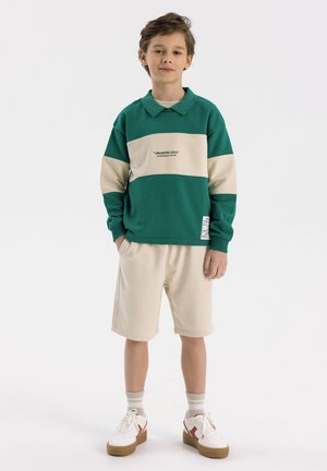 Felpa a blocchi di colore verde e beige con colletto, abbinata a pantaloni corti beige. Presenta un logo stampato e sneakers bianche con accenti rossi.