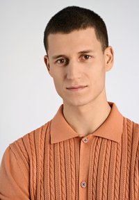 Pfirsichfarbener Strick-Polohemd mit Zopfmuster, kurzen Ärmeln und zwei Knopfverschlüssen am Kragen. Glatte Textur.