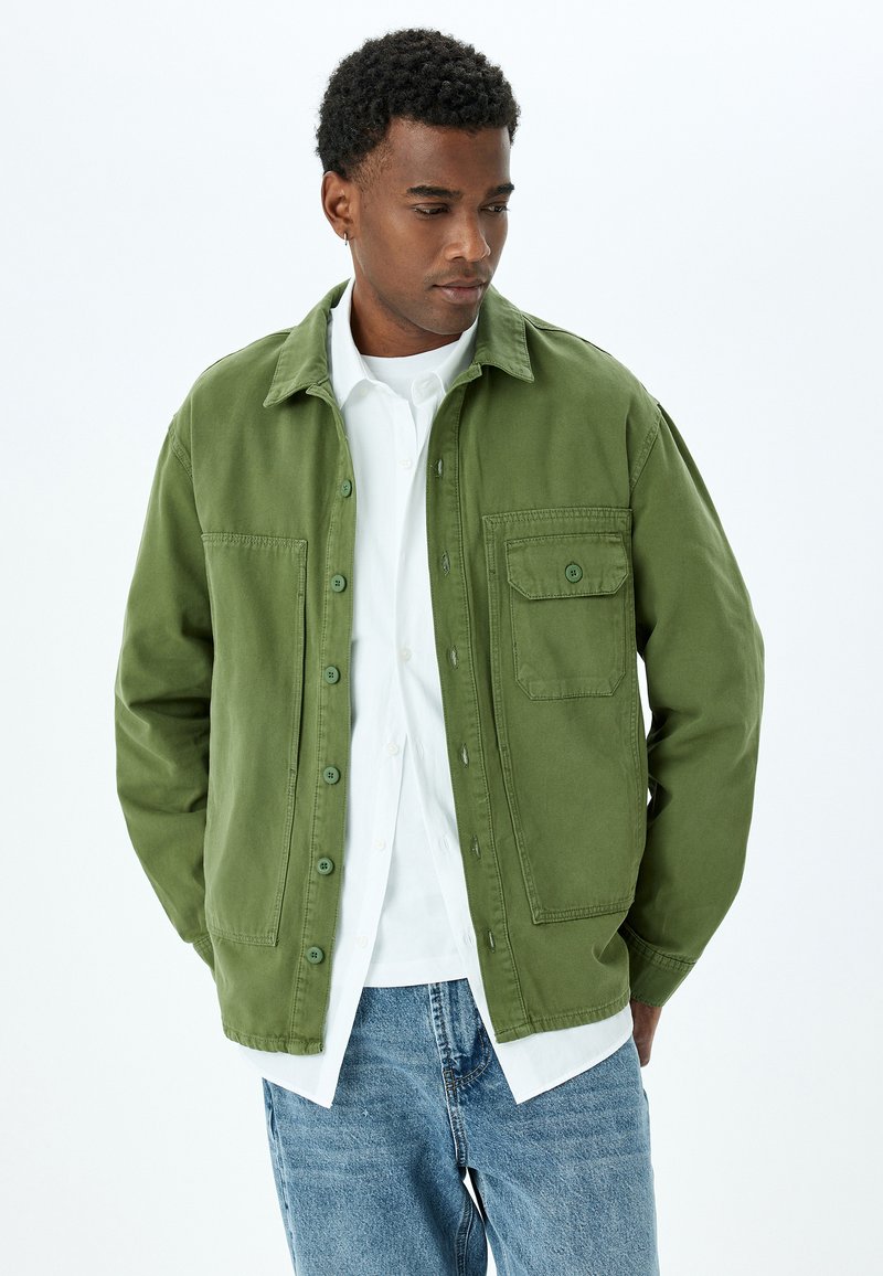 Mies, joka pukeutuu vihreään nappipaitaan valkoisen nappipaidan ja sinisten farkkujen päälle, seisoo kädet taskuissa valkoista taustaa vasten.