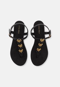 Sandales plates noires avec un design en cœur perlé en or et noir. Comprend des lanières à la cheville et une texture douce semblable à du daim.