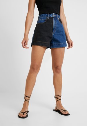Kvinde iført højtaljede shorts med et sort og et blåt denimben, kombineret med sorte sandaler med stropper og en sort ærmeløs top.