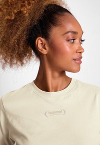 Frau mit lockigem Pferdeschwanz, die ein hellbeiges Hummel Sportswear T-Shirt trägt und auf einen schlichten Hintergrund nach rechts schaut.