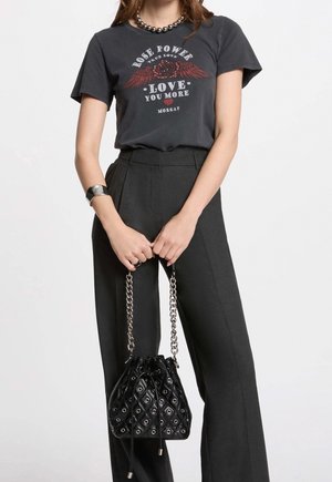 Mujer con camiseta negra gráfica con rosa y texto, pantalones negros de cintura alta, sosteniendo bolso negro acolchado con cadena plateada.