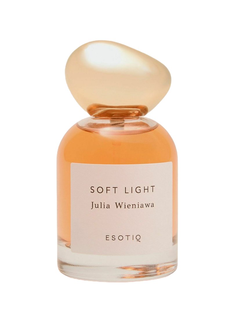 Przezroczysta szklana butelka na perfumy z zaokrągloną bursztynową nakrętką. Zawiera jasno pomarańczowy płyn. Etykieta z napisem „SOFT LIGHT” oraz „Julia Wieniawa.”
