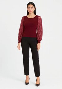 Maglione bordeaux a coste con maniche a sbuffo trasparenti e strutturate, abbinato a pantaloni neri aderenti e tacchi neri, creando una silhouette elegante.