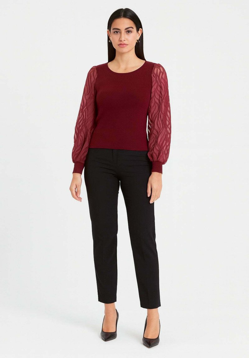 Maglione bordeaux a coste con maniche a sbuffo trasparenti e strutturate, abbinato a pantaloni neri aderenti e tacchi neri, creando una silhouette elegante.