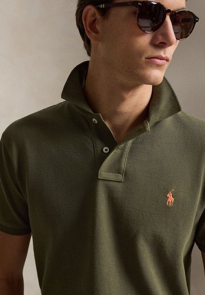 Mann med mørkegrønn poloshorte med oppreist krage og skilpaddemønstrede solbriller, vendt mot høyre.