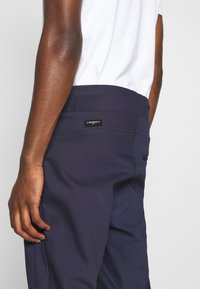 Pantalon bleu marine avec une texture lisse, arborant une étiquette noire sur la taille et des poches latérales. Gros plan sur le bras d'une personne.
