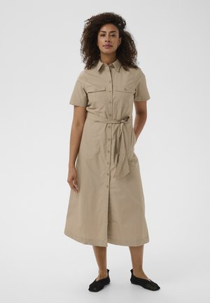 Woman standing wearing a beige button-up midi dress with short sleeves, a waist tie, and black slip-on shoes against a plain background.