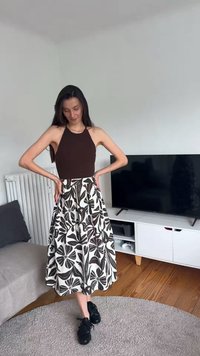 Žena stojící uvnitř s rukama v bok, oblečená v tmavě hnědém topu bez rukávů a bílé sukni s černým květinovým vzorem, černé tenisky.