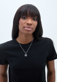 Melna T-kreklu ar īsām piedurknēm, uz kura redzams sudraba kaklarota ar apaļu piekariņu, kurā ir horizontālas līnijas un saules starojuma dizains.