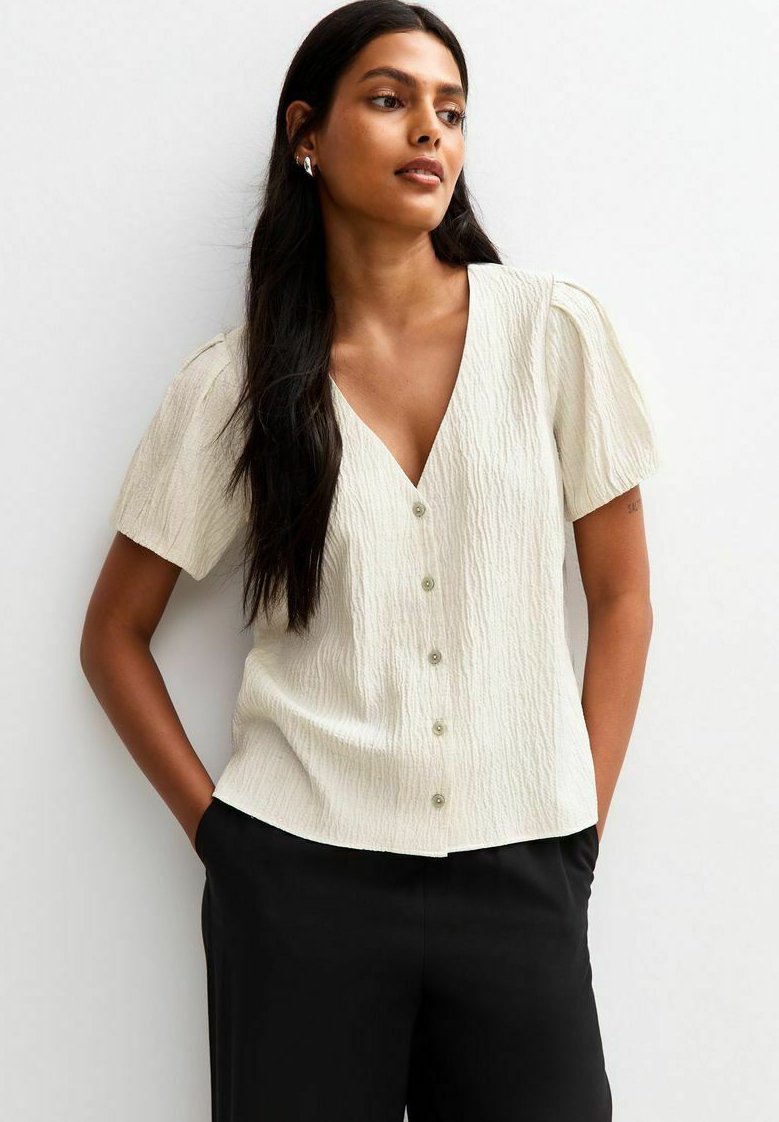 Blusa de manga corta color crema con superficie texturizada, escote en V y cinco botones en la parte delantera. Combinada con pantalones negros.