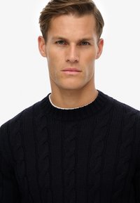 Donkerblauwe kabelgebreide trui met een geribbelde ronde hals en een textuurpatroon, voorzien van een dikke, warme breistructuur.