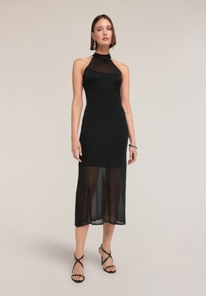 Frau trägt ärmelloses schwarzes Midikleid mit Neckholder und transparentem unteren Rockteil, schwarze Riemchenabsätze und lange Ohrringe, steht vor schlichtem Hintergrund.