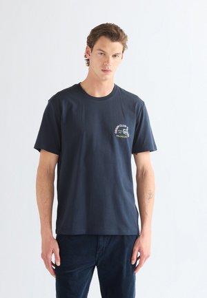 Ung mand med kort brunt hår iført en marineblå T-shirt og mørke bukser, stående foran en ensfarvet lys baggrund.