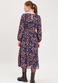 Vestido midi floral con mangas largas, fondo púrpura, patrones azules y beiges, cintura fruncida, espalda con abertura y combinado con botas negras hasta el tobillo.