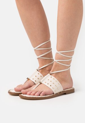 Sandali con lacci caratterizzati da una tomaia sintetica beige con borchie dorate, suola piatta e lunghe fasce avvolte attorno alle caviglie.