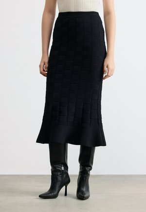 Frau trägt einen schwarzen, strukturierten Midirock mit ausgestelltem Saum und schwarze Leder-Stiefeletten mit hohen Absätzen, steht vor einem einfarbigen Hintergrund.