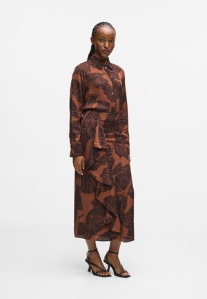 Robe chemise longue marron avec un motif de feuilles noires, boutonnée sur le devant, manches longues, détail de volants asymétriques, associée à des talons noirs à lanières.
