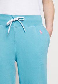 Personne portant un pantalon de survêtement bleu clair avec un cordon blanc noué en nœud et un petit logo rose brodé sur la hanche droite.