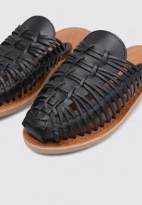 Sandalias de piel trenzada negra con una punta redonda lisa, que cuentan con una plantilla acolchada en color marrón claro y una suela exterior de goma de color claro.