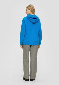 Blauwe hoodie met een ontspannen pasvorm, gecombineerd met zwart-witte houndstooth broek. Soepele textuur en geribbelde manchetten bij de polsen.