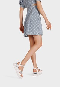 Sandali bianchi con piattaforma, dotati di cinturini regolabili e dettagli metallici, indossati con una gonna di jeans caratterizzata da un motivo geometrico.