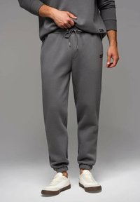 Graue Sweatpants aus weichem Material mit einem elastischen Taillenbund und Kordelzug, gerippten Bündchen und einem kleinen Logo-Patch.