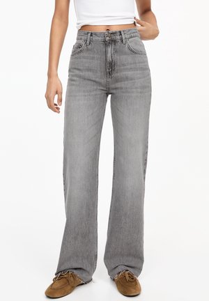 Personne portant un jean gris taille haute à jambes larges, un haut blanc court et des mocassins marron, debout devant un fond blanc uni.