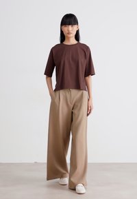Donna con lunghi capelli neri che indossa una maglietta marrone a maniche corte, pantaloni beige larghi, scarpe bianche, in piedi contro uno sfondo chiaro e semplice.