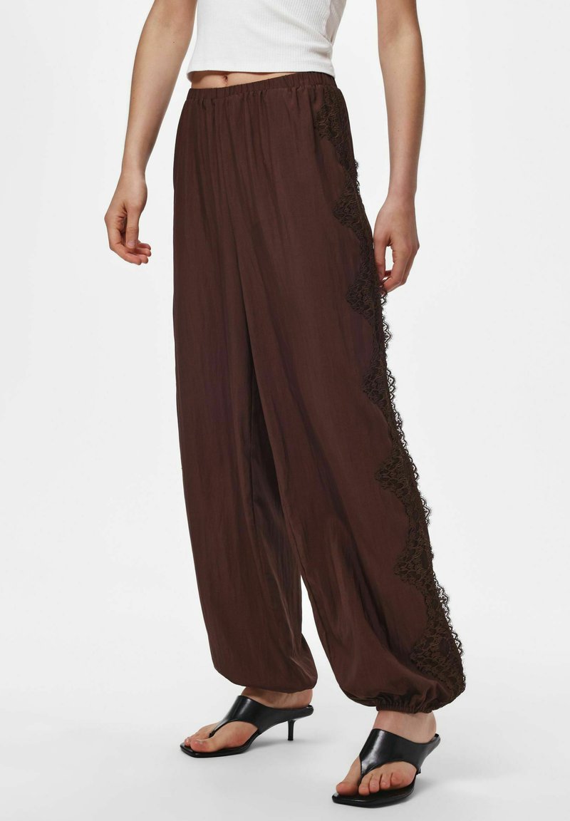 Femme portant un pantalon marron ample avec bordure en dentelle sur les côtés, chevilles élastiquées, débardeur court blanc et sandales à talons ouvertes noires.