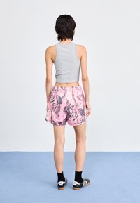 Šedý skrátený top bez rukávov, ružové šortky s listovými vzormi, elastický pás, čierne ponožky, čierno-biele topánky, stojí na modrom pozadí.