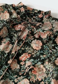 Blouse noire avec motif floral rose et vert, col à volants et détail noué à l'avant.