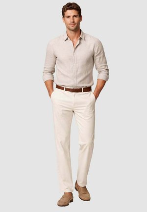 Uomo in piedi con le mani in tasca, indossa una camicia beige a maniche lunghe, pantaloni bianchi, cintura marrone e mocassini in camoscio color cuoio su sfondo chiaro.